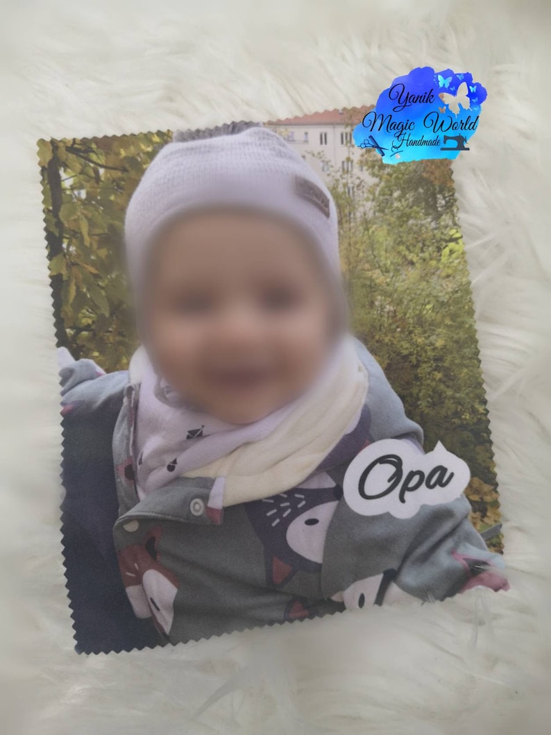 May include: A printed photo of a baby wearing a white hat, a scarf, and a gray jacket with a fox design. The photo has a white label with the word "Opa" on it. The photo is on a white, fluffy surface.