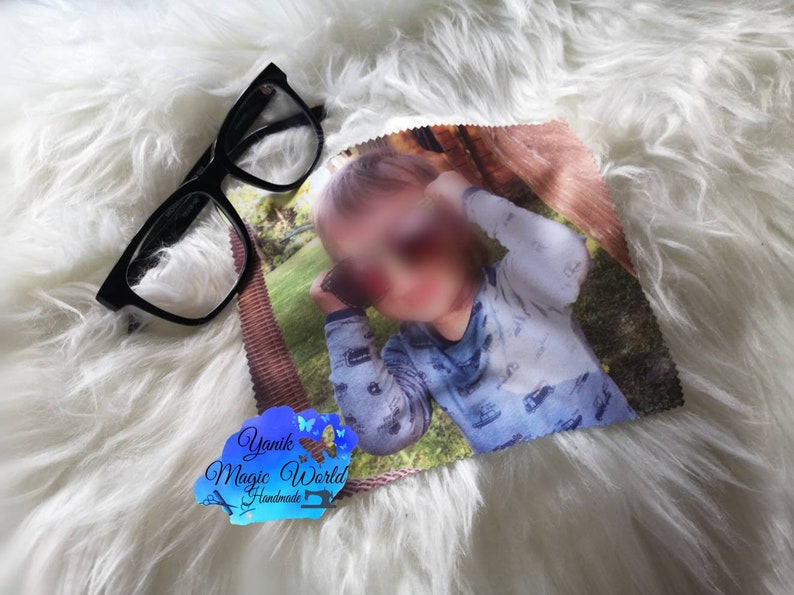 May include: Black-framed eyeglasses and a microfiber cleaning cloth with a printed image of a child. The cloth has a brown border and a tag with the text "Yanik Magic World Handmade". The items rest on a white, fluffy surface.