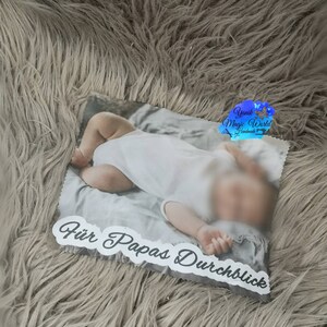May include: A rectangular photo print featuring a baby in a white onesie. The print has the text "F&uuml;r Papas Durchblick" in a decorative font. The background is a soft, fluffy, gray material.