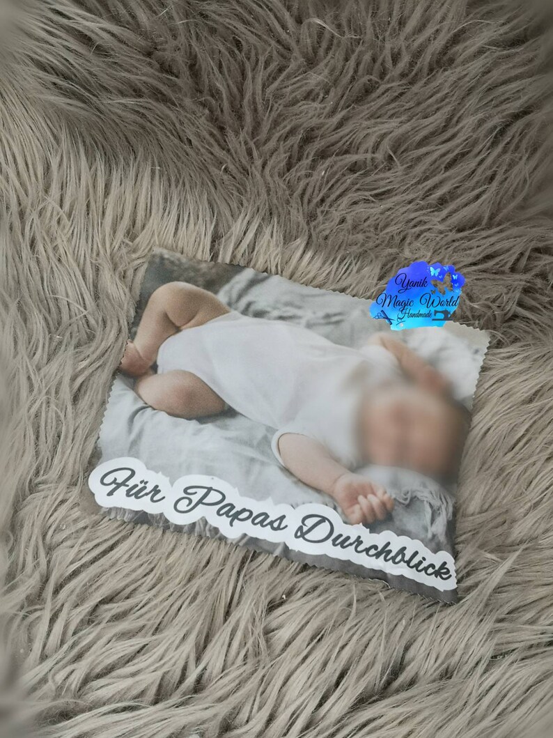 May include: A rectangular photo print featuring a baby in a white onesie. The print has the text "F&uuml;r Papas Durchblick" in a decorative font. The background is a soft, fluffy, gray material.