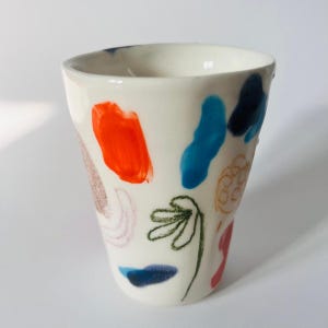 Könnte beinhalten: Ein weißer Keramikbecher mit abstraktem Design. Der Becher hat farbenfrohe Pinselstriche in Orange, Blau und Rot sowie eine grüne Blumenkontur und andere dekorative Elemente. Der Becher hat eine leicht unregelmäßige Form.