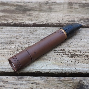 May include: A brown smoking pipe with a black mouthpiece and gold detailing. The cylindrical pipe is made of a dark brown material and has a textured surface. The pipe is resting on a weathered wooden surface.
