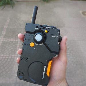 Puede incluir: Un dispositivo IDROID-VER1.02 negro y naranja con una antena elevada, sostenido en una mano. El dispositivo tiene botones etiquetados como "OPEN", "BATTERY", "VOLUME UP/DOWN" y "LIGHT ON-OFF". Tiene un dial circular y pequeños botones.