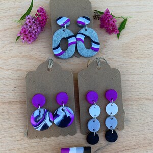 May include: Three pairs of earrings with purple, white, black, and gray polymer clay. The earrings are displayed on brown card stock. A small rectangular pin with purple, white, black, and gray stripes is also shown.