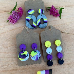 Könnte beinhalten: Drei Paar handgefertigte Ohrringe aus Polymer Clay in verschiedenen Stilen. Das erste Paar zeigt einen großen Oval mit einem grauen Glitzerhintergrund und gelben, violetten und schwarzen Streifen. Das zweite Paar zeigt zwei runde Ohrringe mit einem marmorierten Design in Lila, Gelb und Schwarz. Das dritte Paar zeigt drei runde Ohrringe, die übereinander in Gelb, Weiß, Lila und Schwarz gestapelt sind.