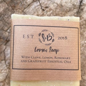 May include: A bar of pale yellow soap with a brown paper label. The label reads "EST S&B 2018 Lemon Soap WITH CLOVE, LEMON, ROSEMARY AND GRAPEFRUIT ESSENTIAL OILS".