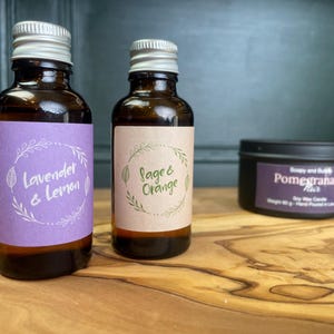May include: Two amber glass bottles with silver screw caps and labels reading Lavender & Lemon and Sage & Orange, alongside a black metal tin labelled Soapy and Bubble Pomegranate Noir Soy Wax Candle with a weight of 80 g printed on the label.