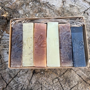 May include: A selection of six rectangular handmade soap bars in a cardboard box. The soaps are in shades of brown, beige, and green. Each bar has an embossed design. The box is presented on a wooden surface.