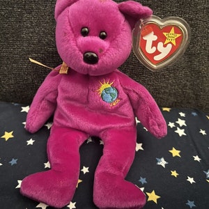 Puede incluir: Peluche de oso Beanie Baby morado con nariz marrón y ojos negros. El oso lleva una etiqueta que dice "Beanie Babies" y tiene un parche en el pecho que dice "Earth 2004".