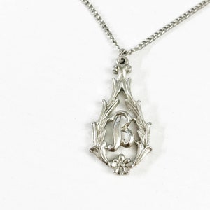 May include: Silver-toned pendant necklace featuring the letter "B" within an ornate, teardrop-shaped frame. The frame has a floral design at the bottom and decorative flourishes. The chain is a delicate curb style, and the background is white.