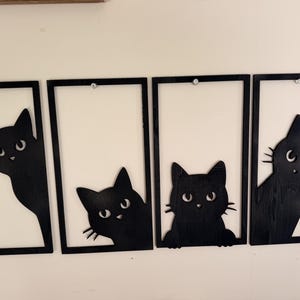 May include: Four rectangular black frames each containing a silhouette of a black cat. The cats are positioned differently in each frame, with some peeking from the sides or bottom, featuring white eyes and whiskers against a plain white background.