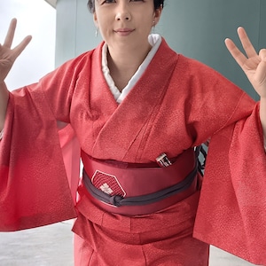 Könnte beinhalten: Eine Frau trägt einen roten Kimono mit einem schwarzen Obi-Gürtel. Sie macht mit beiden Händen das Peace-Zeichen. Der Hintergrund ist eine grüne Wand und ein Parkplatz.