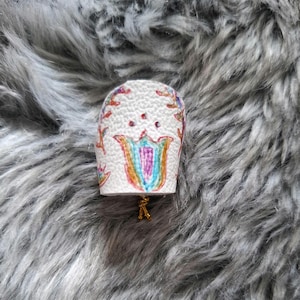 May include: A white, bell-shaped object with a floral design in red, blue, and yellow. The design includes a tulip shape in the center with a gold-colored string attached to the bottom. The object is resting on a gray, furry surface.