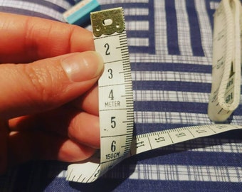 May include: A white measuring tape with black markings, showing measurements in inches and centimeters. The tape is partially extended and shows the numbers 2, 3, 4, 5, and 6. The tape is labeled "150CM" and "METER".