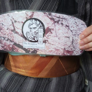 Puede incluir: Una funda rectangular transparente con un diseño de flor de cerezo y el texto "The Ryugiya" e información de la tienda Etsy. La funda se sostiene frente a una persona que lleva un kimono con un estampado de rayas.