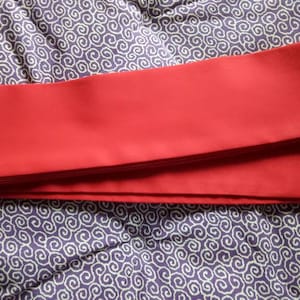 Può includere: Un nastro di stoffa rosso piegato a metà, appoggiato su una superficie con motivo viola e bianco.