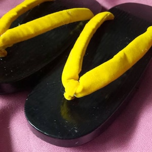 Puede incluir: Un par de sandalias geta negras con correas de tela amarilla. Las sandalias son calzado tradicional japonés.
