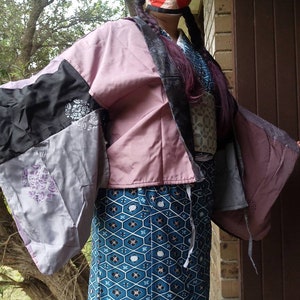 May include: A pink, black and grey patchwork kimono with a blue and white patterned obi. The kimono has a long, flowing sleeve and is tied at the waist with a sash.