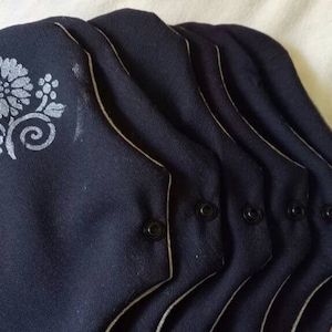 Op de afbeelding: Vijf zwarte herbruikbare stoffen pads met witte bloemenborduursels en witte piping. De pads hebben zwarte drukknopen aan de zijkanten.