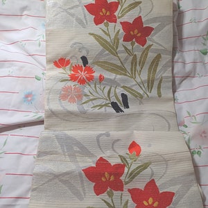 Può includere: Un pannello di tessuto bianco con un bagliore metallico argentato presenta un motivo ripetitivo di fiori rossi e arancioni con foglie verdi. Il tessuto ha una texture a righe sottile.