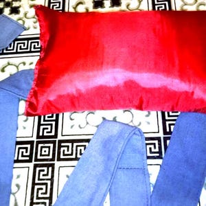 Puede incluir: Una almohada de satén rojo brillante con correas de tela azul. La almohada es rectangular y se encuentra sobre una superficie estampada. Las correas azules están dobladas y cosidas.