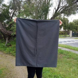 May include: A person is holding a large, dark gray fabric panel in front of them. The fabric is smooth and has a subtle sheen. The person is standing in a grassy area with a house and trees in the background.