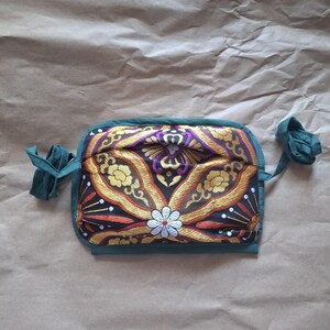 May include: A rectangular fabric pouch featuring a dark background with intricate gold, purple, and orange floral embroidery. The pouch is framed by a solid teal border and includes matching teal fabric ties on both sides for closure.