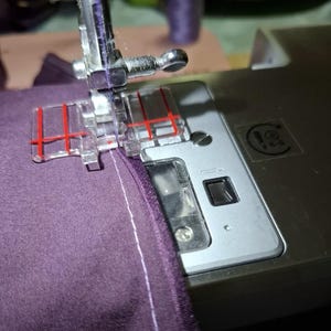 Puede incluir: Primer plano de una aguja de máquina de coser que atraviesa una tela morada. La máquina tiene un pie transparente con marcas rojas.