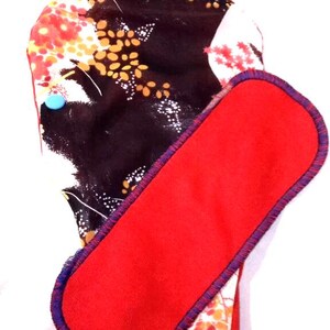 May include: A red cloth menstrual pad with a floral print and a blue trim. The pad is folded in half and has a red fabric insert.