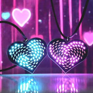 Może przedstawiać: Dwa naszyjniki z wisiorkami w kształcie serca z świecącymi diodami LED. Jeden wisiorek emituje niebieskie światło, drugi różowe. Wisiorki są na czarnych sznurkach, na rozmytym różowo-fioletowym tle.