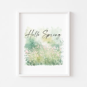 May include: A framed art print with a watercolour painting of a spring meadow. The artwork shows a field of pink flowers and green grass, with the words "Hello Spring" in elegant script. The background has soft green and yellow hues.