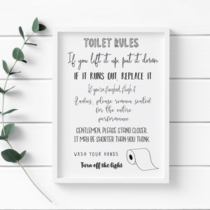 May include: A framed white sign with black text that reads "TOILET RULES" followed by humorous instructions and a drawing of a toilet paper roll. A sprig of green leaves is in the upper left corner.