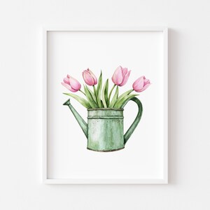 May include: A watercolour painting of a green watering can filled with pink tulips. The tulips have green stems and leaves. The watering can is a light green colour and has a long spout and a curved handle. The artwork is framed in a white frame.