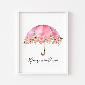 May include: A framed watercolour print featuring a pink umbrella decorated with pink flowers. The umbrella has a wooden handle and the words "Spring is in the air" in black script. The frame is white.