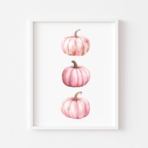 K&ouml;nnte beinhalten: Gerahmter Kunstdruck mit drei rosa Aquarellk&uuml;rbissen auf wei&szlig;em Hintergrund. Jeder K&uuml;rbis hat einen braunen Stiel. Die vertikale Anordnung ist schlicht und elegant, ideal f&uuml;r die Herbstdekoration.