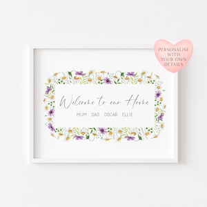 May include: A white framed print featuring the words "Welcome to our Home" within a floral border of daisies and purple flowers. The names "MUM DAD OSCAR ELLIE" are below the text. A pink heart graphic is in the top right corner.