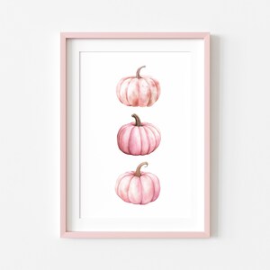 K&ouml;nnte beinhalten: Gerahmter Kunstdruck mit drei rosa Aquarellk&uuml;rbissen auf wei&szlig;em Hintergrund. Die K&uuml;rbisse haben braune Stiele. Der hellrosa Rahmen erg&auml;nzt die Illustration und schafft eine herbstliche Atmosph&auml;re.