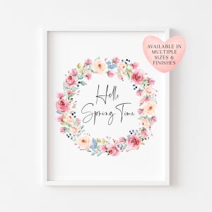 May include: A rectangular white frame containing a print with a floral wreath of pink and cream roses and green leaves surrounding the black text Hello Spring Time. A pink heart-shaped graphic in the top right corner reads AVAILABLE IN MULTIPLE SIZES & FINISHES.