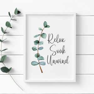 Puede incluir: Una impresi&oacute;n enmarcada en blanco con las palabras "Relax Soak Unwind" en escritura negra. La impresi&oacute;n presenta una rama de eucalipto en acuarela con hojas verdes y verde azulado. El fondo es una pared de paneles de madera blanca.