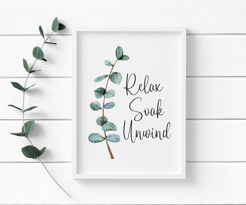 Puede incluir: Una impresi&oacute;n enmarcada en blanco con las palabras "Relax Soak Unwind" en escritura negra. La impresi&oacute;n presenta una rama de eucalipto en acuarela con hojas verdes y verde azulado. El fondo es una pared de paneles de madera blanca.
