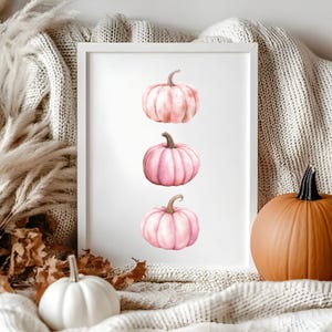 K&ouml;nnte beinhalten: Ein gerahmter Druck mit drei rosa K&uuml;rbissen mit braunen Stielen. Die K&uuml;rbisse sind vertikal auf wei&szlig;em Hintergrund angeordnet. Ein wei&szlig;er K&uuml;rbis und ein gro&szlig;er orangefarbener K&uuml;rbis befinden sich im Vordergrund, zusammen mit einer neutral get&ouml;nten Decke und getrocknetem Laub.