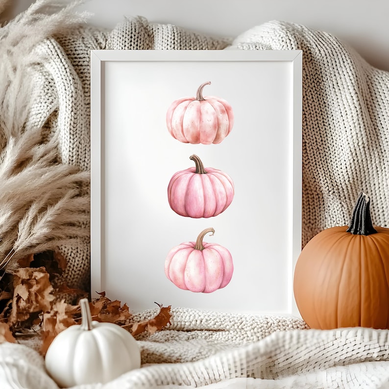 K&ouml;nnte beinhalten: Ein gerahmter Druck mit drei rosa K&uuml;rbissen mit braunen Stielen. Die K&uuml;rbisse sind vertikal auf wei&szlig;em Hintergrund angeordnet. Ein wei&szlig;er K&uuml;rbis und ein gro&szlig;er orangefarbener K&uuml;rbis befinden sich im Vordergrund, zusammen mit einer neutral get&ouml;nten Decke und getrocknetem Laub.