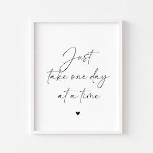 May include: A framed print featuring the phrase "Just take one day at a time" in black cursive script. A small black heart is at the bottom. The print is in a white frame, ideal for interior decoration.