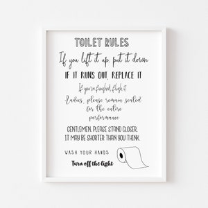 May include: A framed print with the words "TOILET RULES" in black script. The rules are listed below, including "If you lift it up, put it down" and "Turn off the light." A roll of toilet paper is illustrated.