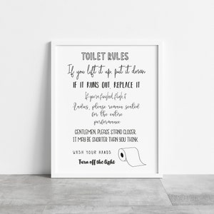 May include: A framed print with the text "TOILET RULES" in a black, handwritten-style font. The rules include instructions like "If you lift it up, put it down" and "Wash your hands." A simple illustration of a toilet paper roll is included.