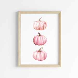 K&ouml;nnte beinhalten: Gerahmter Druck mit drei rosa Aquarellk&uuml;rbissen. Jeder K&uuml;rbis hat einen Stiel und ist vor einem wei&szlig;en Hintergrund platziert. Die K&uuml;rbisse sind vertikal angeordnet und bilden eine einfache, elegante Komposition.