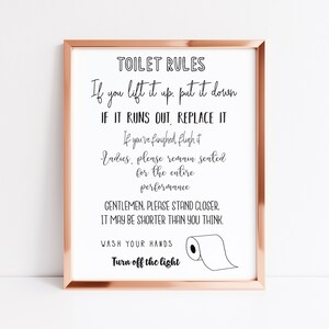 May include: A framed print with a rose gold border featuring the text "TOILET RULES" in black script. The rules include instructions like "If you lift it up, put it down" and "Wash your hands." A simple illustration of a toilet paper roll is included.