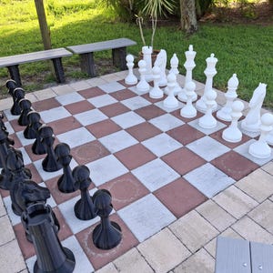 May include: An outdoor chess set with large, black and white pieces arranged on a red and white checkered board. The chess pieces are set up on a patio, ready for a game. Two benches are visible in the background.
