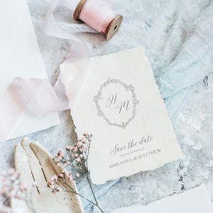 May include: A white card with a floral wreath and the initials "A & M" in the center. The text "Save the date for the wedding of Adrianna & Matthew" is printed below the wreath.