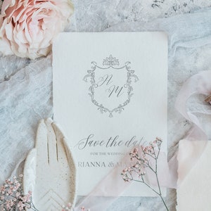 K&ouml;nnte beinhalten: Save-the-Date-Karte mit einem zarten floralen Design und einem Monogramm der Buchstaben "R" und "M" in einem dekorativen Rahmen. Der Text lautet "Save the date for the wedding of Rianna & Matthew".